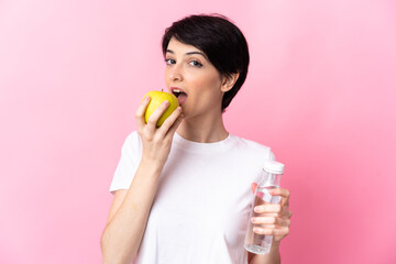 Woman with short hair isolated on pink background with a bottle of water and eating an apple