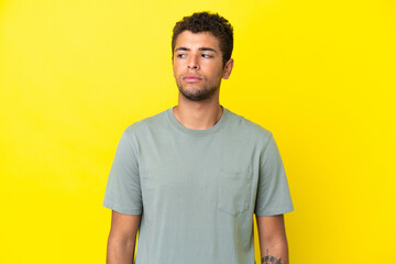 Fototapeta premium Young handsome Brazilian man isolated on yellow background having doubts while looking side