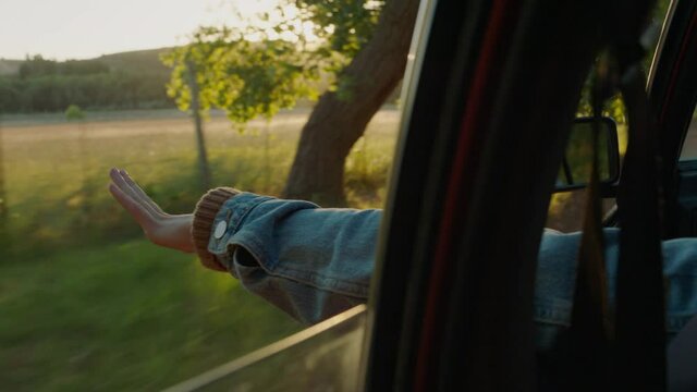 woman in car holding hand out window feeling wind blowing through fingers driving in countryside travelling on summer vacation road trip enjoying freedom on the road at sunset