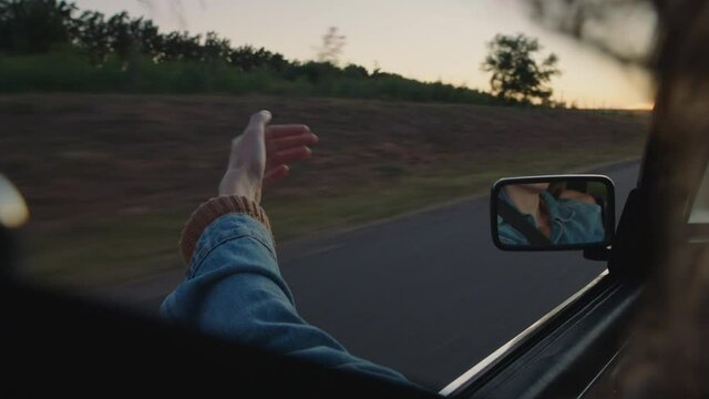 young woman in car holding hand out window feeling wind blowing through fingers driving in countryside on road trip travelling for summer vacation enjoying freedom on the road at sunset