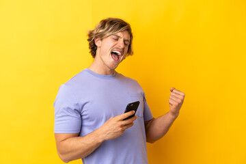 Handsome blonde man isolated on yellow background with phone in victory position