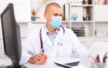 Man doctor in medical mask is working at laptop in clinic office