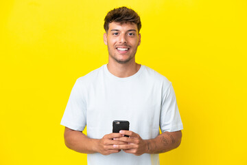 Young caucasian handsome man isolated on yellow background looking at the camera and smiling while using the mobile