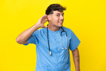 Young surgeon doctor man isolated on yellow background listening to something by putting hand on the ear