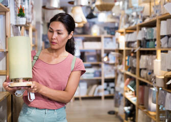 Asian woman choosing table lamp in furniture store. She's holding one in hands.