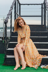A girl in long dress sitting with bare feet on the stairs on the top of the roof of building.