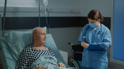 Nurse checking file of sick patient laying in hospital ward bed at recovery unit talking about treatment for healing pain and disease. Elder man with nasal oxygen tube and oximeter