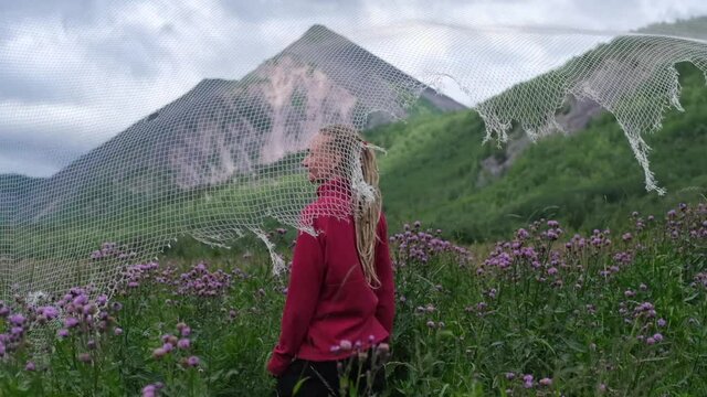 Slow motion video portrait of a young woman standing on a summer meadow. Grass swaying in the wind, mountain landscape, soccer goal net, football field in the village