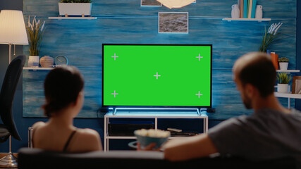 Young man and woman watching green screen on tv display sitting on couch at home. Caucasian people...