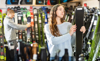 Fototapeta na wymiar Portrait of young smiling woman choosing new skis in shop of sports equipment