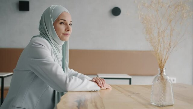 Portrait of beautiful young woman in elegant hijab turning to camera and smiling indoors in halal cafe at table. Islam religion and people concept.