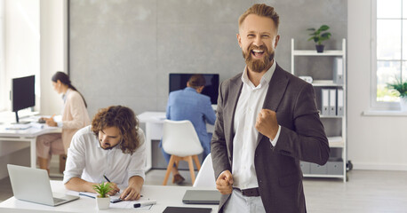 Portrait of handsome man fist pumping with happy face expression standing in modern office with people working in background. Businessman manager wins business deal, finds solution, celebrates success