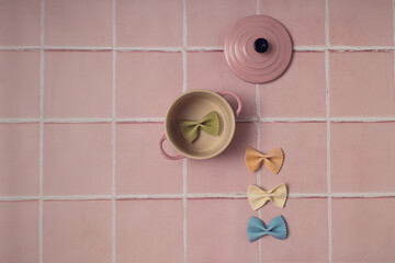 Raw uncooked Italian pasta colorful bow tie farfalle in a pink casserole on a pink tile kitchen counter top. Top view. Food background with copy space. Flat lay with negative space.
