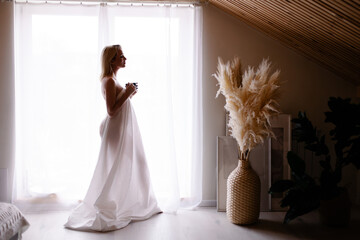 Early awakening morning magic. woman in sheet stands by the window drinking coffee from mug. Slow...