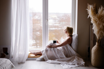 Early awakening morning magic. woman in sheet stands by the window drinking coffee from mug. Slow down leisure lifestyle. Start of the weekend mood. Date with yourself. Slowdown. Digital detox. autumn