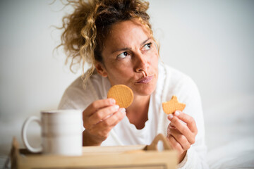 Worried adult female sith biscuits and coffee in morning breakfast at home laying on bed - pretty middle age woman with food and nutrition thoughts and problems for weight