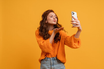 Attractive red head woman in stylish fall outfitm sweater and jeans posing over yellow  background in studio.   Girl sen kiss to smartphone . © Svetlana Sokolova