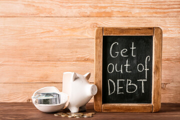 Chalkboard with text GET OUT OF DEBT, piggy bank and money on wooden background