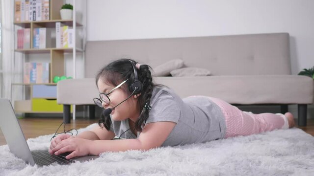 Happy Asian girl wearing glasses, lying on the floor, taking video call on laptop and using wireless microphone in living room, typing on keyboard during having video call. Staying home concept