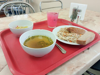 On the table there is a tray with broth with meatballs, pearl barley porridge with chicken cutlet, salad with tomatoes and cucumbers, jelly with cranberries and black bread
