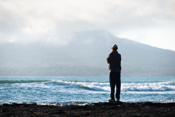 Man standing on Milford beach and watching the waves rolling in, with out-of-focus Rangitoto Island in the mist.