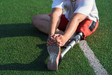 Close up of a Caucasian guy with a prosthetic leg stretching while sitting on the field at the stadium. Sport concept.