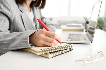 Young woman with laptop writing something in notebook at home