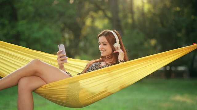 Young beautiful red-haired woman resting on hammock. Cheerful girl wear headphones having fun. Pretty lady enjoys listening chill music