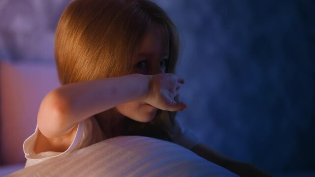 Upset blonde little girl with dimly lit face leaning on soft pillow wipes running nose and tears on bed near wall in semidark bedroom at moon light slow motion