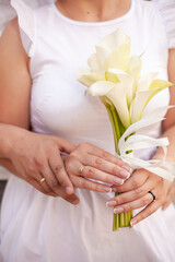 The bride is holding a beautiful wedding bouquet of lilies in her hands. The groom hugs from behind. Close-up photo. Touch of hands