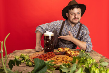 Happy smiling man dressed in traditional Austrian or Bavarian costume sitting at table with festive food and beer isolated over red background