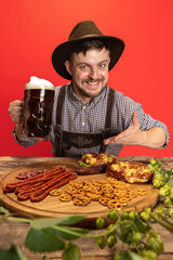 Happy smiling man dressed in traditional Austrian or Bavarian costume sitting at table with festive food and beer isolated over red background