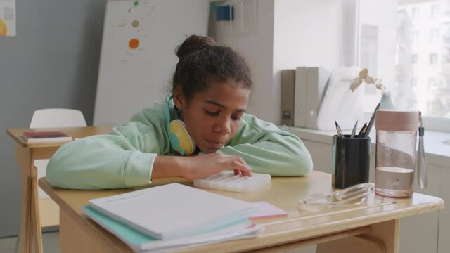 Handheld shot of African-American 11-year-old girl with headphones sitting at desk in classroom and playing with sensory fidget toy