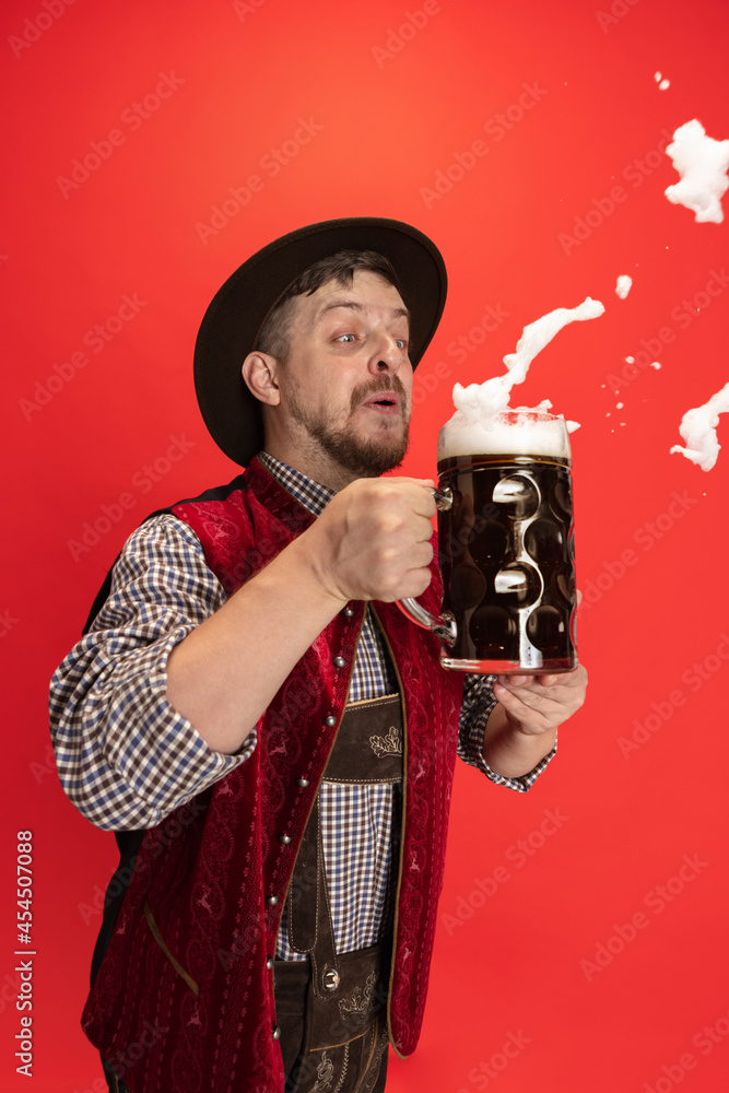 Wall mural Happy smiling man dressed in traditional Austrian or Bavarian costume holding mug, glass of cold foamed dark black beer isolated over red background