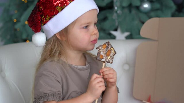 4k video portrait of one cute little child eating Christmas chocolate sweets happily. Craft chocolate handmade sweets in hands of kid. Pretty blonde little girl wearing red holiday Santa hat
