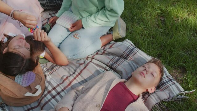 Handheld tracking shot of children relaxing on blanket laid out on green grass in park. Girls are chatting and playing with sensory fidget toys while boy lying with his eyes closed