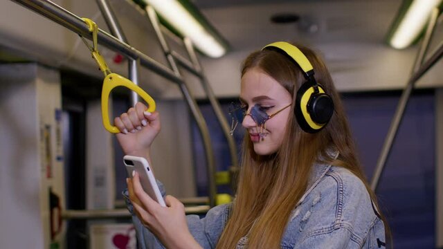 Young woman wearing headphones listening music, funny relax dancing while traveling by bus to city center. Citizen. Public transport. Urban lifestyle. Attractive girl passenger at modern tram or train