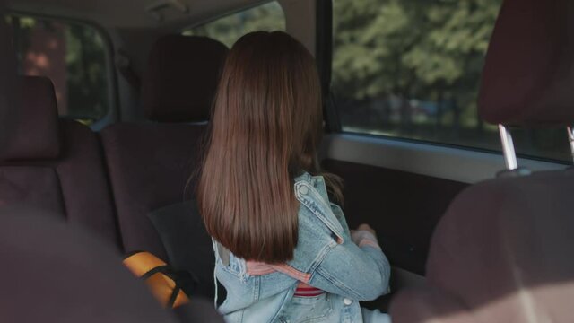 Shot from inside of car cheerful 12-year-old girl with backpack getting into passenger seat of car and fastening seatbelt, the smiling to person off camera