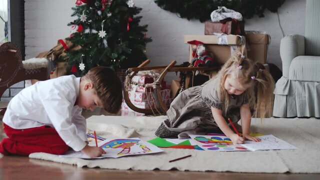 Closeup view 4k video footage of 2 cute little kids coloring special black and white pictures in coloring books using colorful bright pencils. Small boy and girl sitting on floor in christmas interior