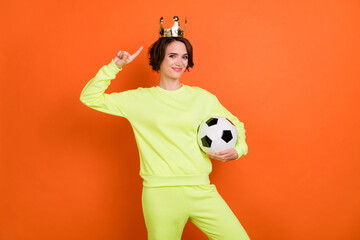 Portrait of trendy beautiful cheery girl holding in hand ball demonstrating tiara isolated over bright orange color background