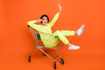 Portrait of attractive cheerful funny girl riding cart showing v-sign rest isolated over bright orange color background