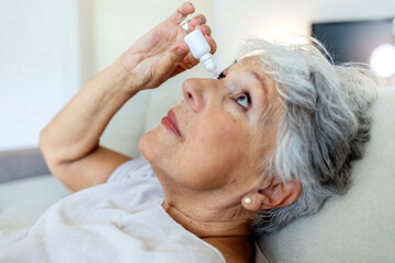 Naklejka premium Cropped shot of a elderly female person using eye drops. Photo of a senior woman putting eye drop, close up view of an elderly person using a bottle of eyedrops in her eyes. Concept of eye treatment.