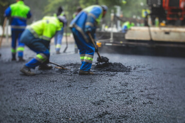 Process of asphalting, blacktopping and paving, group of workers with shovels finishing the asphalt layer, with asphalt paver machine and steam roller compactor vehicle in the background