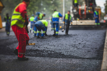 Process of asphalting, blacktopping and paving, group of workers with shovels finishing the asphalt layer, with asphalt paver machine and steam roller compactor vehicle in the background