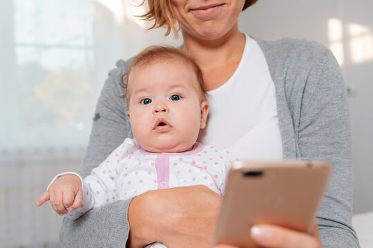 Portrait of a baby sitting in the arms of a smiling mother who uses a smartphone. Bottom view. Smartphone is blurred in the foreground