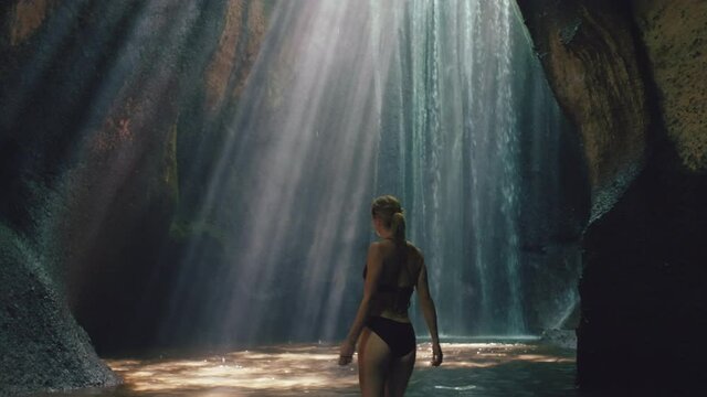beautiful woman in waterfall cave wearing bikini exploring underground cavern alone with water splashing through light rays