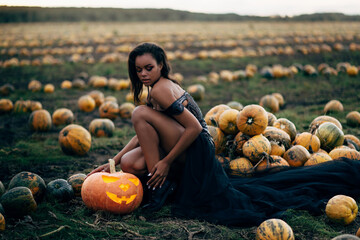 Young black woman sitting near scary face pumpkin in field. Halloween concept.