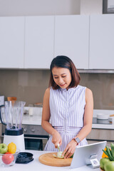 Asian woman cutting vegetables in kitchen with tablet in front of her