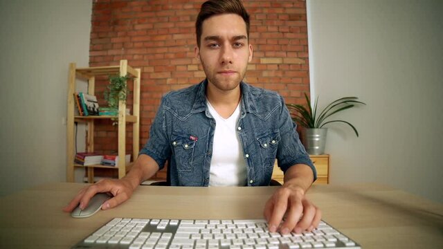 POV of focused handsome male freelancer working at desktop computer, typing on wireless keyboard and using mouse, looking at camera. Webcam view of concentrated young man working on pc at home office.
