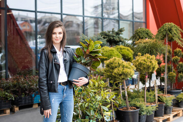 Fototapeta na wymiar A young pretty girl, dressed in a leather jacket and jeans, stands near the flower center and holds a large beautiful flowerpot.